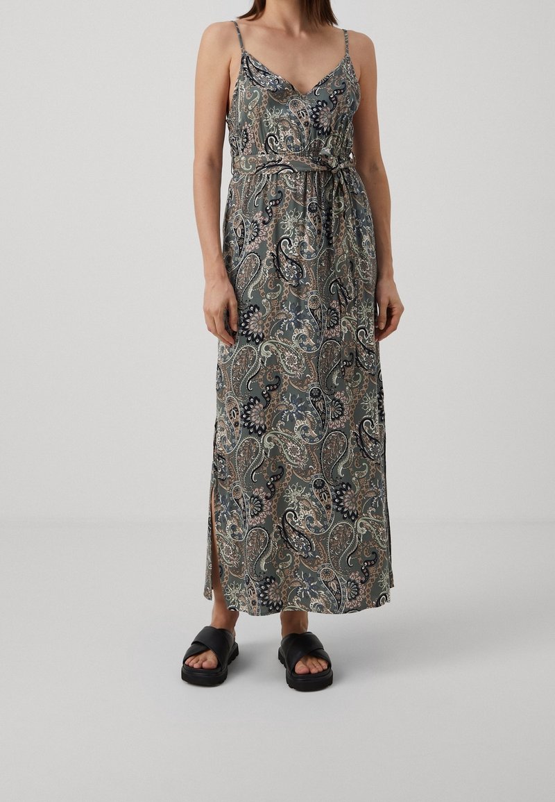 Femme portant une robe longue sans manches à motif cachemire, avec des fentes latérales et des sandales noires à enfiler, sur un fond uni.