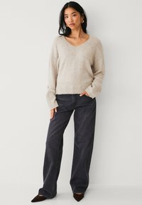 Beige gebreide trui met een V-hals en ribbelrand, gecombineerd met donkere wijduitlopende jeans en puntige bruine schoenen. Glad materiaal en ontspannen pasvorm.