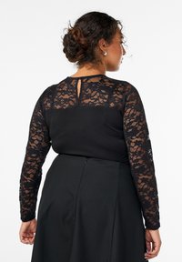 Ženska s temnimi lasmi spet v bunki, oblečena v črno obleko z čipkastimi dolgimi rokavi in zgornjim delom hrbta, stoji obrnjena proti beli ozadju.