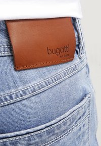 Vaqueros de mezclilla azul claro con una etiqueta de cuero marrón en la cinturilla con las inscripciones "bugatti" y "est. 1978" en relieve. Se ven dos bolsillos traseros.