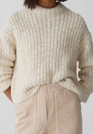 Frau trägt einen grob gestrickten, cremefarbenen Pullover mit Rippenstruktur und beige hoch taillierte Cordhosen, die Hände in den Taschen vergraben.