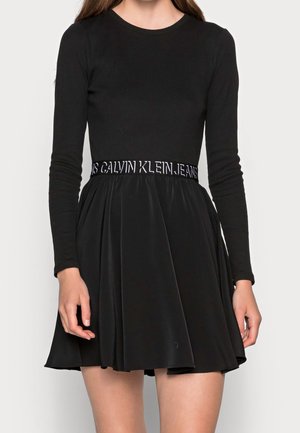 Frau trägt ein eng anliegendes schwarzes Langarmshirt und einen schwarzen ausgestellten Rock mit der Aufschrift "Calvin Klein Jeans" auf dem Taillenbund.