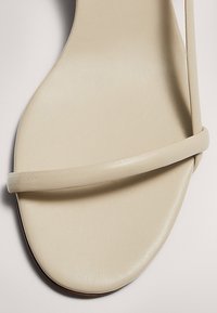 Sandale minimaliste beige avec une fine lanière à l'avant et un design à bout carré sur un fond neutre.