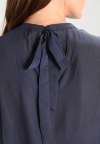 Nära bild av en kvinnas rygg som visar en marinblå blus med en knuten rosett i nacken och en mjuk tygtextur.