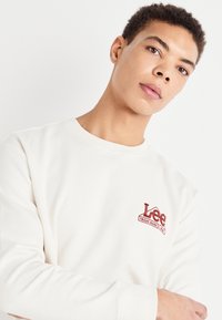 Felpa color crema con scollo rotondo e maniche lunghe, con logo "Lee" ricamato in rosso sul petto. Texture liscia.