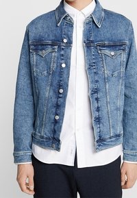 Džínová bunda světle modré barvy s tmavým prošíváním, obsahující dvě náprsní kapsy, stříbrné kovové knoflíky a klasický design límce.