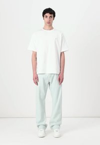 Camiseta de algodón blanca de manga corta combinada con pantalones de corte relajado verde claro y zapatillas blancas, de pie contra un fondo liso.