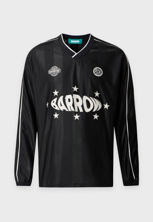 Joven de pie con una camiseta negra de manga larga con texto blanco y vaqueros anchos azules con gráficos coloridos y zapatos tipo slip-on a cuadros.