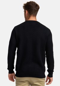 Marineblauwe trui met een gestructureerd chevronpatroon, ronde halslijn en geribde manchetten en zoom; gedragen over lichtgekleurde broek.