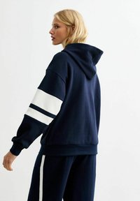 Marine-blauwe hoodie met witte streepaccenten op de mouwen, voorzien van een voorzak en een trekkoordcapuchon. Zachte stof met een relaxed fit.