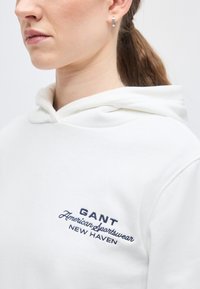 Femme portant un sweat à capuche blanc avec l'inscription "GANT American Sportswear New Haven" brodée sur la poitrine, petit anneau d'argent visible à l'oreille.