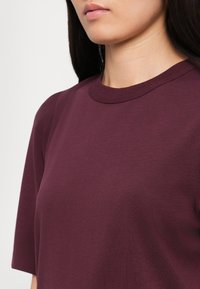 Gros plan d'une personne portant un t-shirt uni à manches courtes, col rond, de couleur bordeaux foncé, montrant le cou et le bas du visage.