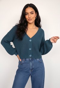 Miętowy dzianinowy kardigan z głębokim dekoltem w kształcie litery V, pięcioma niebieskimi guzikami i długimi rękawami. Połączony z wysokim stanem niebieskich dżinsów.