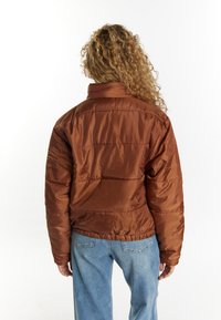 Brun isoleret puffjakke med høj krave, elastisk talje og horisontale quilte detaljer. Båret over lyseblå denimjeans.