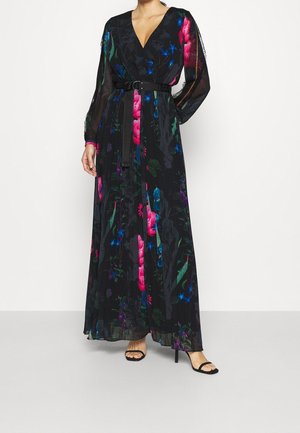 Kobieta ubrana w długą czarną sukienkę w kwiatowy wzór z prześwitującymi rękawami i paskiem w talii, w połączeniu z czarnymi wysokimi butami na otwartym palcu.