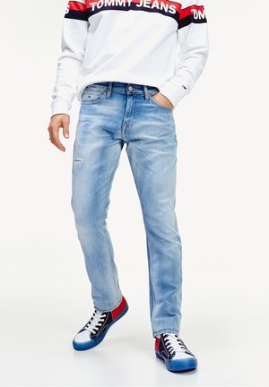 Personne portant un jean bleu clair, un haut blanc à manches longues avec le logo Tommy Jeans rouge et marine, et des baskets noires, blanches, rouges et bleues.