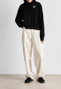 Haut noir court avec une poche avant et un logo blanc, associé à un pantalon beige clair ample ; des baskets noires complètent la tenue.