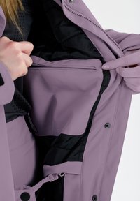 Interno di una giacca impermeabile lilla che mostra un rivestimento nero e una tasca con zip, dotata di chiusure con cordino e bottoni a pressione. Texture liscia.