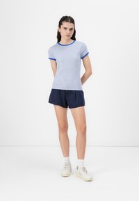 Ριγέ μπλε και άσπρη μπλούζα με μπλε φινίρισμα, σε συνδυασμό με ναυτικές βερμούδες. Το μοντέλο φοράει λευκά αθλητικά παπούτσια και κάλτσες. Απλός σχεδιασμός, casual εφαρμογή.