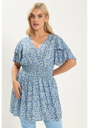 Lyshåret kvinde iført en blå blomsterprintet V-hals kjole med flagrende ærmer og smocktalje, stående foran en ensfarvet lys baggrund.