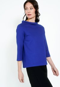 Kobaltkék, háromnegyedes ujjú felső, cseppnyak kialakítással, puha anyagból készült, laza szabású és sima textúrájú.