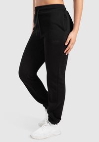 Zwarte joggers van zacht materiaal met een trekkoord in de taille, zijzakken en geribde boorden. Kenmerkt een subtiele logo-detail.