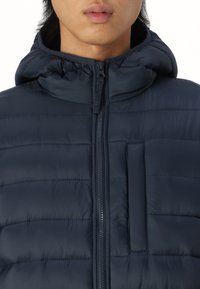 Navy blå puffjakke med glidelås, hette og horisontale sømmer; har en brystlomme for ekstra funksjonalitet.