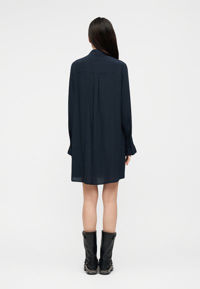 Γυναίκα με μακριά σκούρα μαλλιά στέκεται με την πλάτη, φορώντας ένα χαλαρό σκούρο μπλε φόρεμα με μακριά μανίκια και μαύρες μπότες μέχρι τη μέση της γάμπας με αλυσιδωτές λεπτομέρειες.