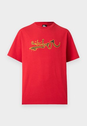 Jeune homme portant un t-shirt rouge oversize avec un design graphique abstrait et un short en jean bleu clair, debout devant un fond uni.