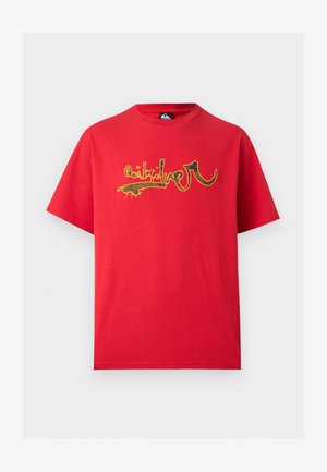 Mladý muž v červenom oversize tričku s abstraktným grafickým dizajnom a svetlomodrých denimových krátkych nohaviciach stojaci na jednoliatom pozadí.