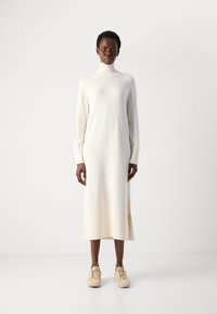 Femme se tenant debout, portant une longue robe-pull blanche à col roulé avec des fentes sur les côtés et des chaussures beiges à lacets sur un fond uni.