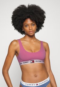 Sujetador deportivo rosa con una amplia banda gris que presenta el logo de "TOMMY HILFIGER". Con tirantes gruesos y textura suave, combinado con ropa interior azul.