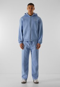 Ljusblå zip-up hoodie med kängurufickor, matchad med ljusblå byxor som har sidofickor. Tyget verkar mjukt och slätt.