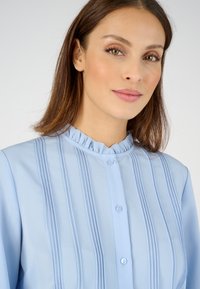 Blusa de color azul claro con cuello con volantes, cierre de botones y pliegues verticales. La tela suave y la textura ligera proporcionan un aspecto limpio y pulido.