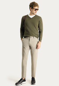 Suéter verde oliva con escote en forma de V, pantalones beige, zapatillas negras con suelas blancas, correa de hombro marrón y gafas de sol. Tejido suave con un diseño ajustado.