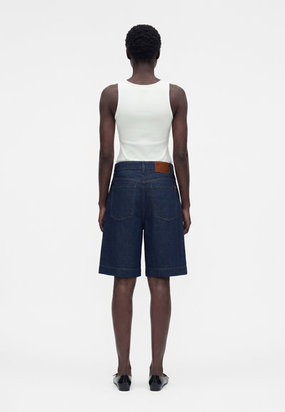 Άτομο με κοντά σγουρά μαλλιά που φοράει λευκό αμάνικο τοπ, σκούρα μπλε τζιν σορτς μέχρι το γόνατο και μαύρα παπούτσια, στέκεται γυρισμένο προς τα πίσω.