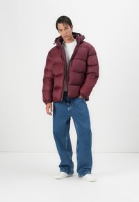 Chaqueta de plumas burdeos con capucha, presenta secciones acolchadas, puños elásticos y una cremallera delantera. Lleva puesta una camiseta gris y jeans azules.