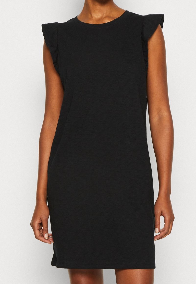 Woman wearing a sleeveless black dress with ruffled shoulders, standing against a plain light background.