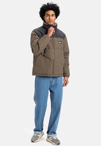Pufferjack in olijfgroen en zwart, met een hoge kraag, ritssluiting en zijzakken, gecombineerd met lichtblauwe jeans en sneakers.