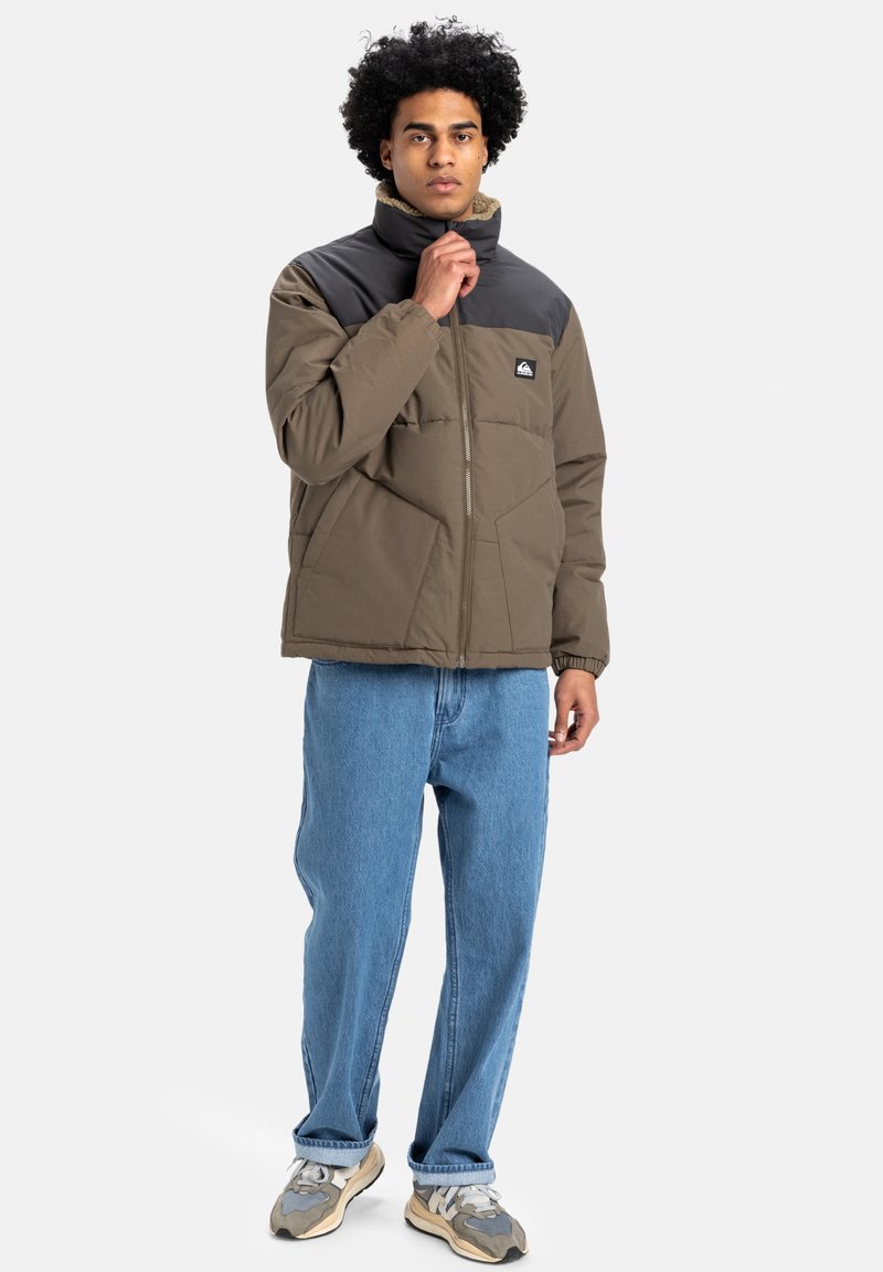 Pufferjack in olijfgroen en zwart, met een hoge kraag, ritssluiting en zijzakken, gecombineerd met lichtblauwe jeans en sneakers.