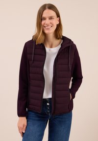 Femme souriante portant une veste à capuche violette foncée sur une chemise blanche et un jean bleu, se tenant contre un fond uni.