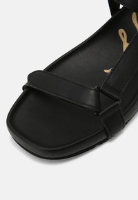 Sandale plate noire avec larges brides sur les orteils et la cheville, dotée d'une semelle coussinée et de détails de couture minimalistes.