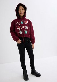 Felpa con cappuccio color bordeaux con ricami festivi di regali, fiocchi di neve e bastoncini di zucchero; abbinata a pantaloni neri aderenti e stivali neri.