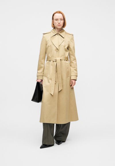 Άτομο που φοράει μπεζ καμπαρντίνα με ζώνη, σκούρο παντελόνι με φαρδιές μπατζάκια, μαύρα μυτερά παπούτσια, διάφανα γυαλιά και κρατά μαύρη τσάντα χειρός σε λευκό φόντο.