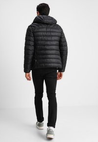 Mężczyzna w czarnej pikowanej kurtce z kapturem, czarnych dżinsach i szarych trampkach odchodzi na białym tle.