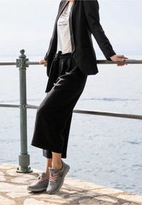 Chaussures de sport grises avec des lacets roses, portées avec un pantalon noir raccourci et un t-shirt blanc à imprimé, sur un fond de bord de mer.