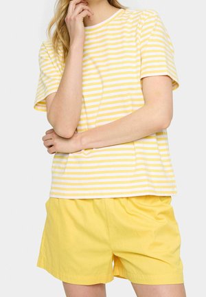Frau trägt ein gelb-weiß gestreiftes kurzärmliges Shirt mit einfarbigen gelben Shorts, posiert mit einer Hand nahe am Gesicht und einem verschränkten Arm.