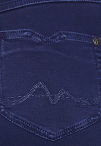 Mörkblå denimtyg har ett distinkt vågigt sömmönster på bakfickan, kompletterat med en cirkulär metalldetalj.