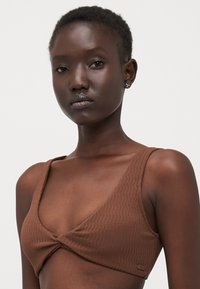 Brun teksturert bralette med vridd frontdesign, brede stropper og en glatt finish. Har en subtil logo på siden.