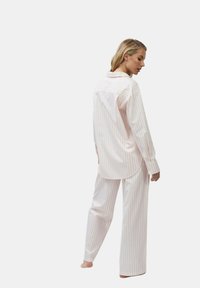 Frau steht barfuß, trägt ein weißes Langarmshirt und eine Hose mit feinen vertikalen roten Streifen und schaut nach links vor einem schlichten Hintergrund.